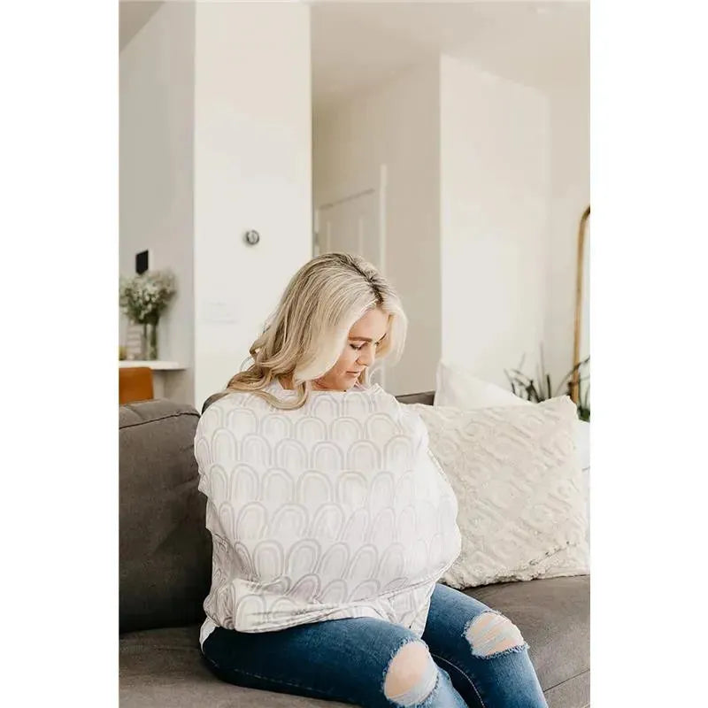 A woman seated on a couch uses a Bliss Canopy baby car seat cover by Copper Pearl, depicted in a modern living space.