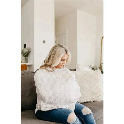 A woman seated on a couch uses a Bliss Canopy baby car seat cover by Copper Pearl, depicted in a modern living space.