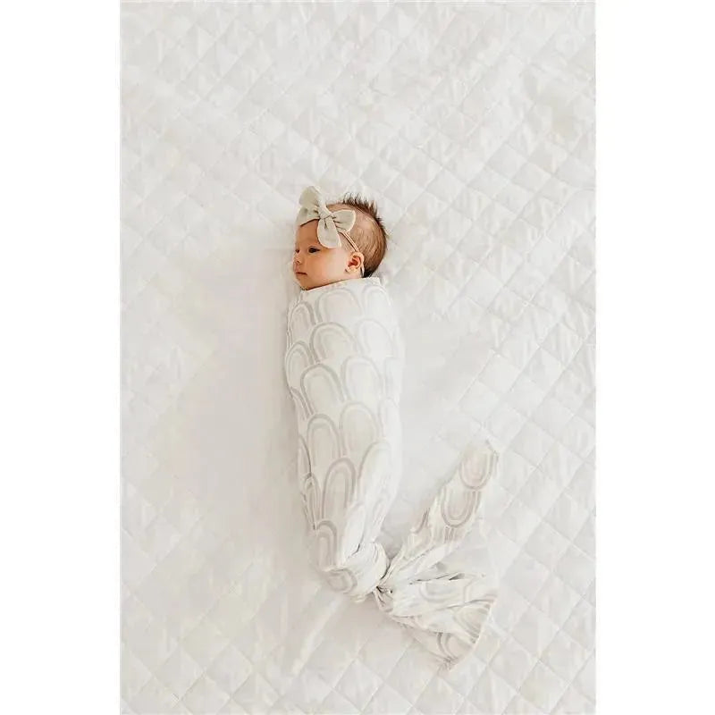 Bliss Knit Baby Swaddle from Copper Pearl featuring a baby wrapped in a neutral-colored blanket with a bow headband.