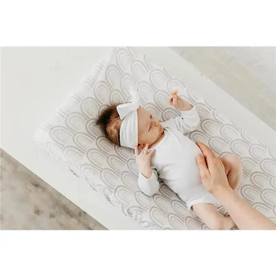 A baby wearing a white onesie and headband lies on a Copper Pearl Bliss Knit diaper changing pad cover with a rainbow design.