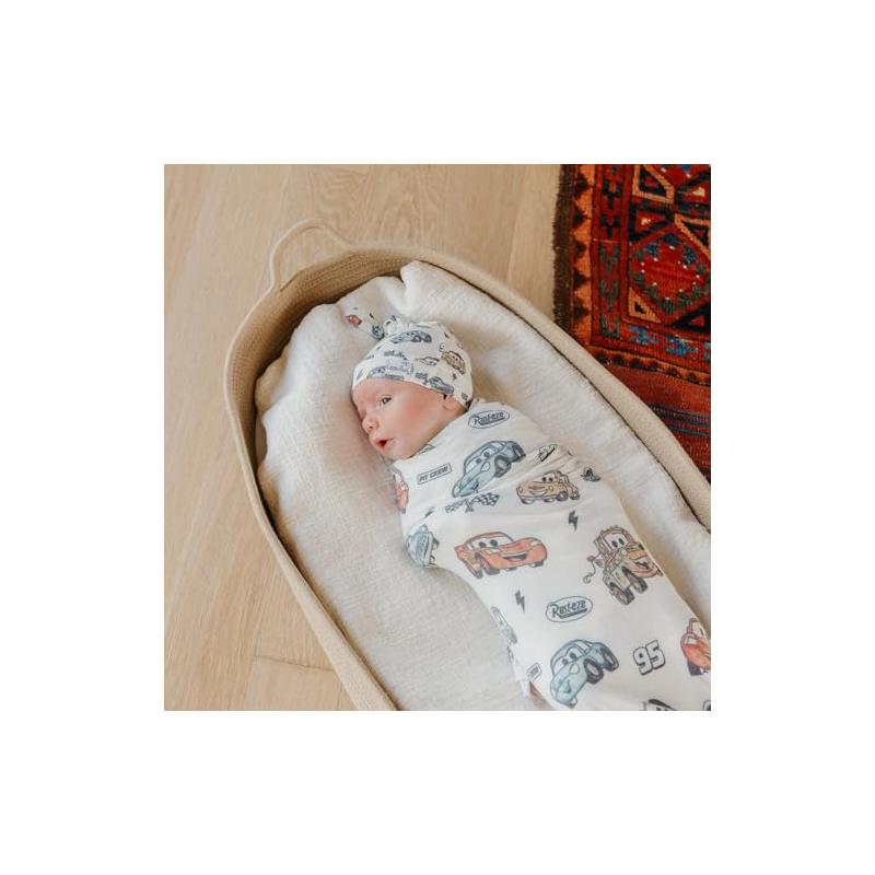 Infant wrapped in a swaddle featuring Disney Pixar Cars characters, wearing a matching top knot hat, relaxing in a bassinet.