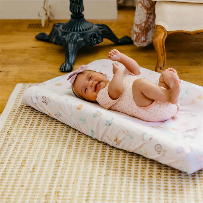 Baby lying on a Copper Pearl Enchanted Friends diaper changing pad cover, smiling with a bow headband, on a woven rug.