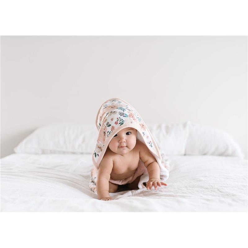 Infant wearing a floral-printed hooded towel, crawling on a white bedspread.