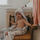 Baby sitting on a chair wearing a Copper Pearl Hunny Premium Knit Hooded Towel, smiling with the hood up.