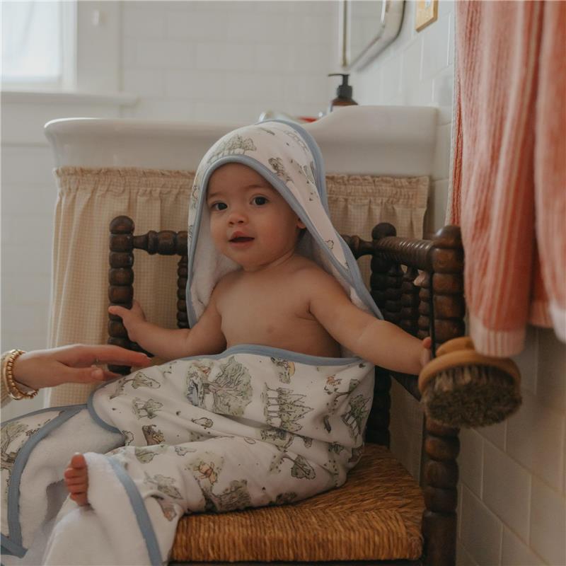 Infant wrapped in a Copper Pearl Hunny Premium Knit Hooded Towel with cartoon designs, sitting on a wooden chair in a bathroom.