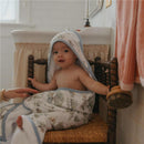 Infant wrapped in a Copper Pearl Hunny Premium Knit Hooded Towel with cartoon designs, sitting on a wooden chair in a bathroom.