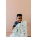 Young child smiling while holding a Copper Pearl Rex Knit Baby Swaddle Receiving Blanket with a leafy pattern.