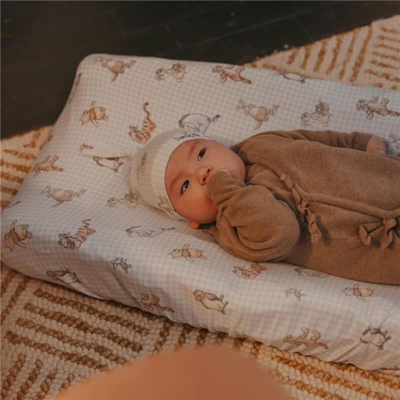 A baby lies on a Copper Pearl Winnie The Pooh diaper changing pad cover, wearing a soft hat and a brown outfit.