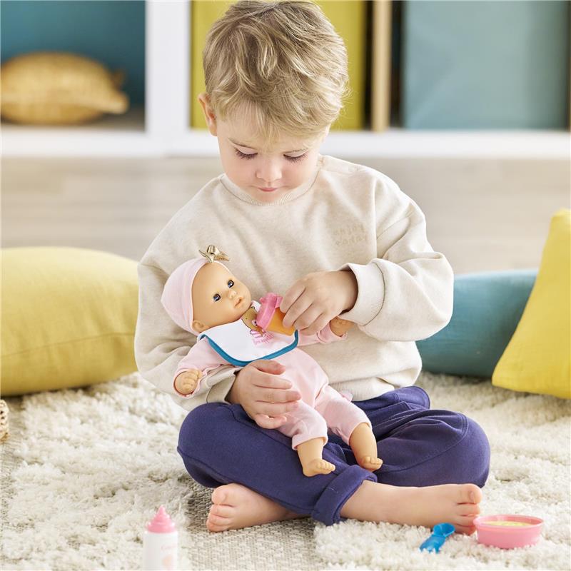 Child in casual clothing feeding a Corolle baby doll with a bottle, surrounded by colorful cushions and a soft area rug.