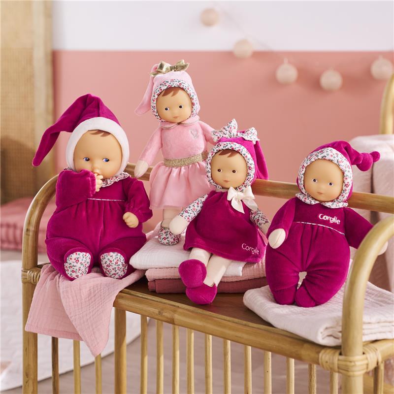Four Corolle Babipouce dolls dressed in pink and purple outfits are displayed on a wooden shelf with soft blankets.