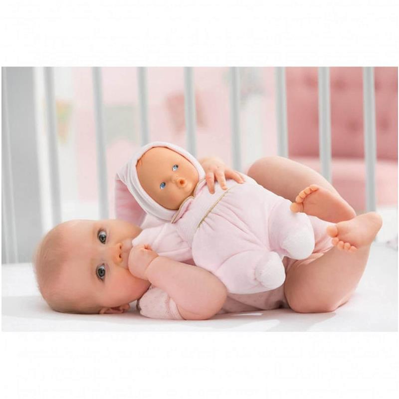 A baby holds a Corolle Babipouce Sweet Dreams soft doll in pink attire while lying on a white crib mattress.
