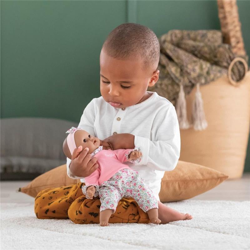 A child sits on a patterned pillow, gently cradling a Corolle Bébé Calin Maria doll wearing a pink shirt and floral pants.