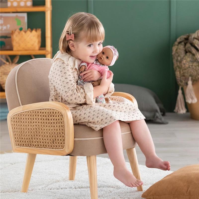 A young girl in a patterned dress sits on a wooden chair, holding a Corolle Bébé Calin Maria baby doll with a pink hat.
