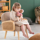 A young girl in a patterned dress sits on a wooden chair, holding a Corolle Bébé Calin Maria baby doll with a pink hat.