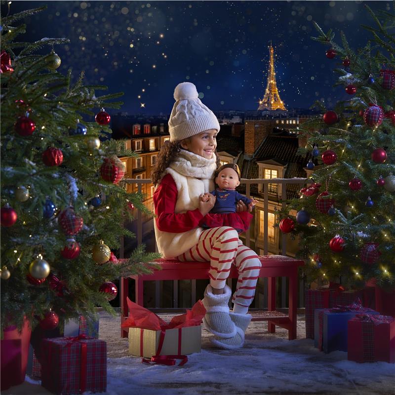 Girl in winter attire sitting on a red bench holds a Corolle Ma Perle doll, with decorated Christmas trees and the Eiffel Tower in the background.