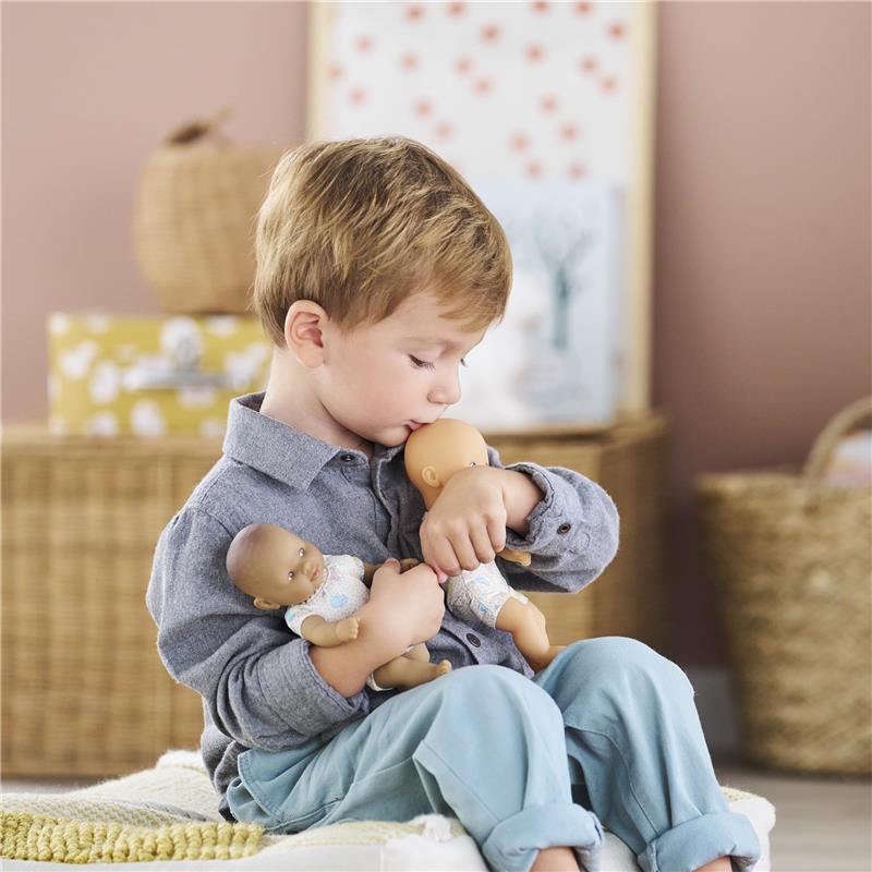 A young boy in a gray shirt sits, gently kissing a blonde baby doll while holding a second doll in his lap.