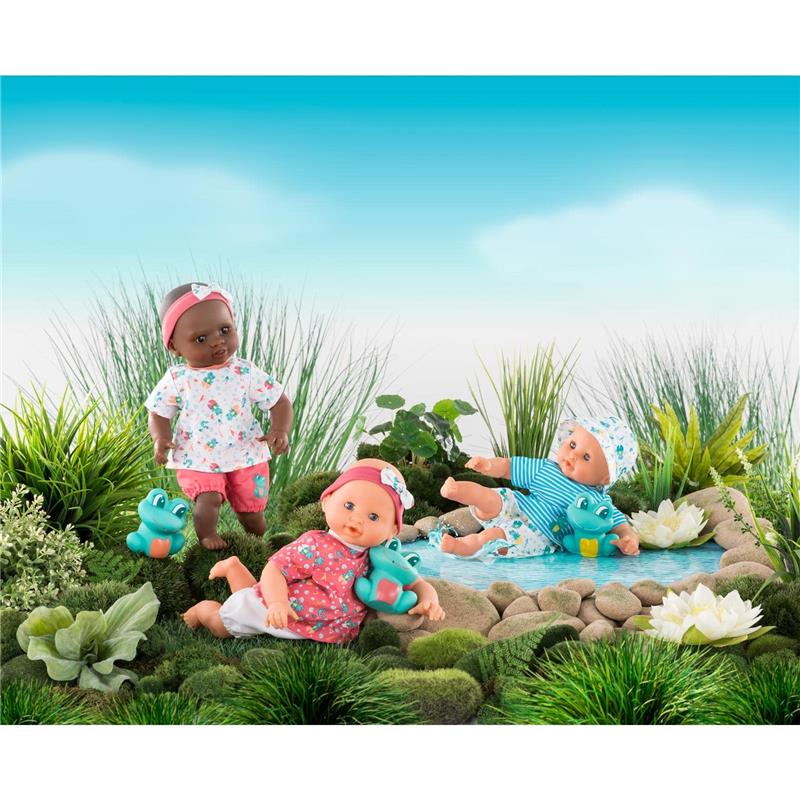 Three Corolle dolls in colorful outfits playing by a small pond, with one doll holding a frog toy.