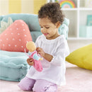 Young girl in a white blouse kneels while holding a Corolle My First Doll, dressed in a pink outfit, in a colorful playroom.