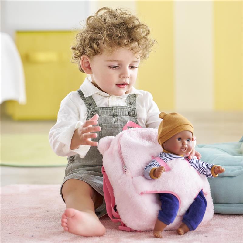 Child playing with a pink plush backpack baby doll carrier, holding a doll dressed in a striped shirt and beanie.