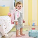 Child wearing a gray overall stands with a plush pink backpack that carries a baby doll, smiling in a colorful bedroom.