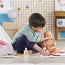 Child using a toothbrush on a Corolle doll, with accessories including a pink bag, lotion bottle, and glass.