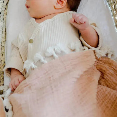 Baby lying on a soft surface, partially wrapped in a copper red muslin swaddle blanket, wearing a light-colored shirt.