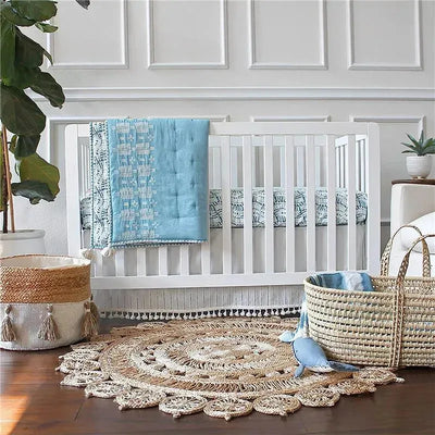 White crib with a blue quilt draped over it, surrounded by a cozy nursery setting with woven baskets and a circular rug.