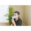 A young man is using the Crane cordless warm and cool mist inhaler, visibly inhaling mist while seated indoors.