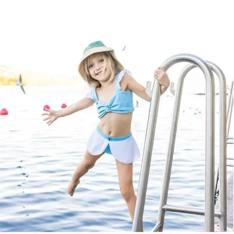 A young girl in a blue and white two-piece swimsuit, smiling while climbing a pool ladder near water.