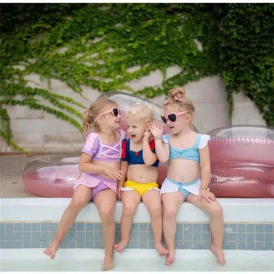 Three young girls in colorful swimsuits sit on the edge of a pool, smiling and playing together, surrounded by floaties.