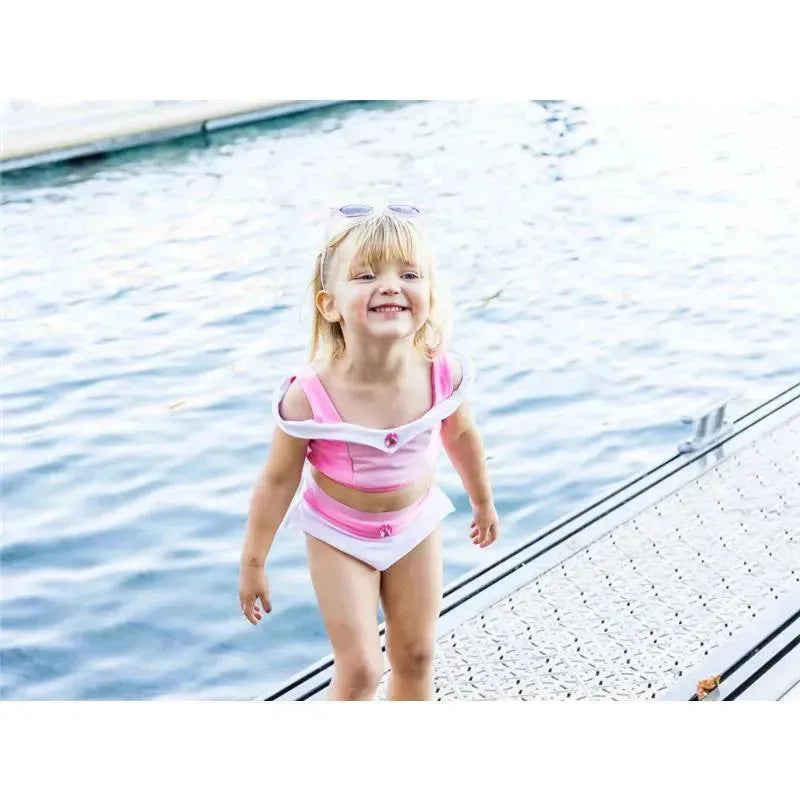 Young child in a pink swim suit with white accents, smiling and playing near water on a dock.