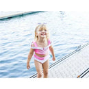Young child in a pink swim suit with white accents, smiling and playing near water on a dock.
