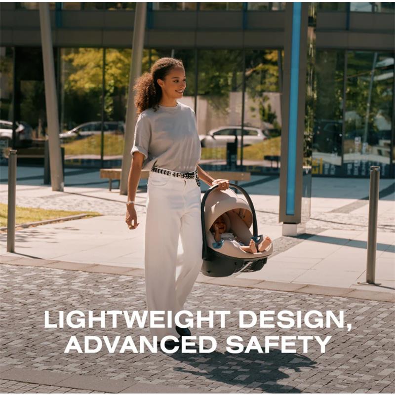 A woman walks outdoors, carrying a Cybex Aton G2 swivel infant car seat in almond beige, with a smiling baby inside.