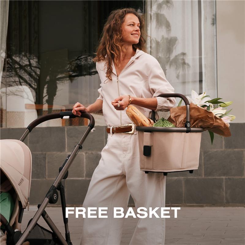 A woman smiles while holding a stroller and a cream-colored basket filled with groceries and flowers.