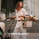 A woman smiles while holding a stroller and a cream-colored basket filled with groceries and flowers.