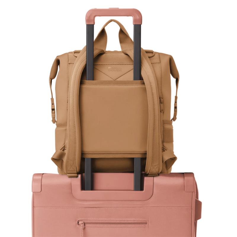 Large Dagne Dover Indi Diaper Backpack in camel with backpack straps and a slot for a luggage handle, resting on a pink suitcase.
