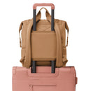 Large Dagne Dover Indi Diaper Backpack in camel with backpack straps and a slot for a luggage handle, resting on a pink suitcase.