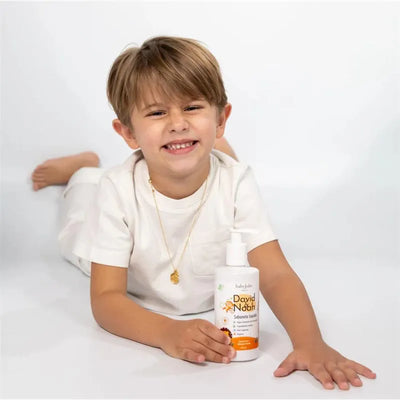 Boy in a white shirt smiles while lying down, holding a bottle of David & Noah By Baby Jolie body wash.