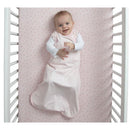 Smiling baby in a pink and white striped sleep sack with built-in swaddle, lying in a crib with a pink dotted sheet.