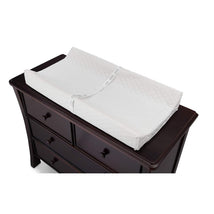 Contoured changing pad on a dark wood dresser, featuring a quilted white cover and safety strap for secure use.