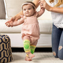 Green and yellow dinosaur knee pads on an infant wearing a pink romper, assisted by an adult while standing.