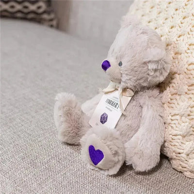 Gray plush bear with purple heart and nose, labeled as February Birthstone, sitting on a textured surface.