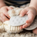A baby’s hand grasps a gray, textured teething toy alongside a soft, plush giraffe attachment.