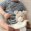 Demdaco Giraffe Teether Buddy is a plush teether featuring a giraffe head with a soft beige and white knit body, held by a seated baby in a striped outfit.