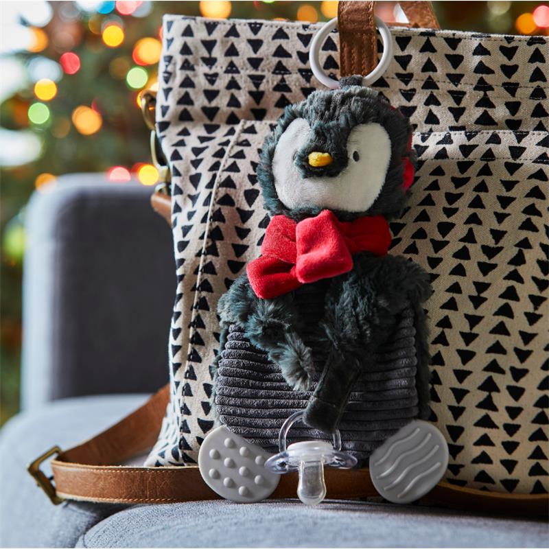 Penguin-shaped teether buddy with a red bow, attached to a patterned bag, set against a blurred festive background.