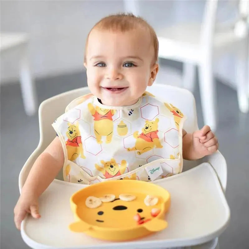 Toddler wearing a Winnie the Pooh bib sits in a high chair, smiling at a silicone Pooh-themed dish with sliced bananas.