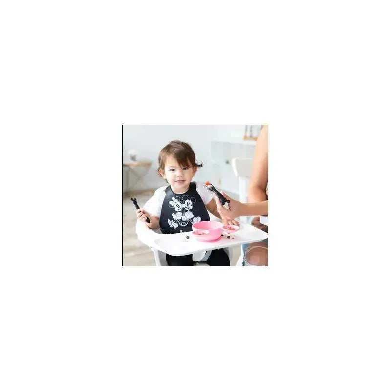 Child in a high chair wearing a Mickey Mouse bib, holding silicone dipping spoons with a pink bowl in front.