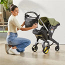 A woman in a light sweater kneels beside a Doona stroller, attaching a black Essentials Bag. The stroller features a green canopy.