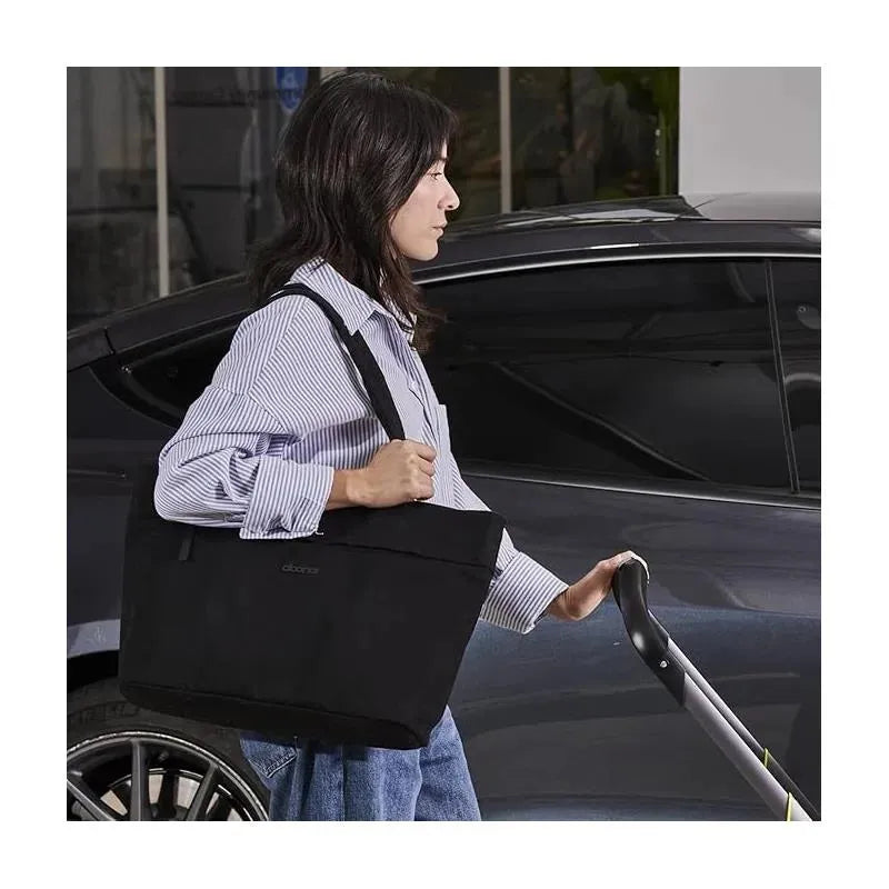 A woman carrying a black Doona Essentials Diaper Tote Bag while holding the handle of a stroller near a car.