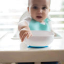 Baby reaching for a white no-slip suction bowl on a high chair tray, with a blurred background.